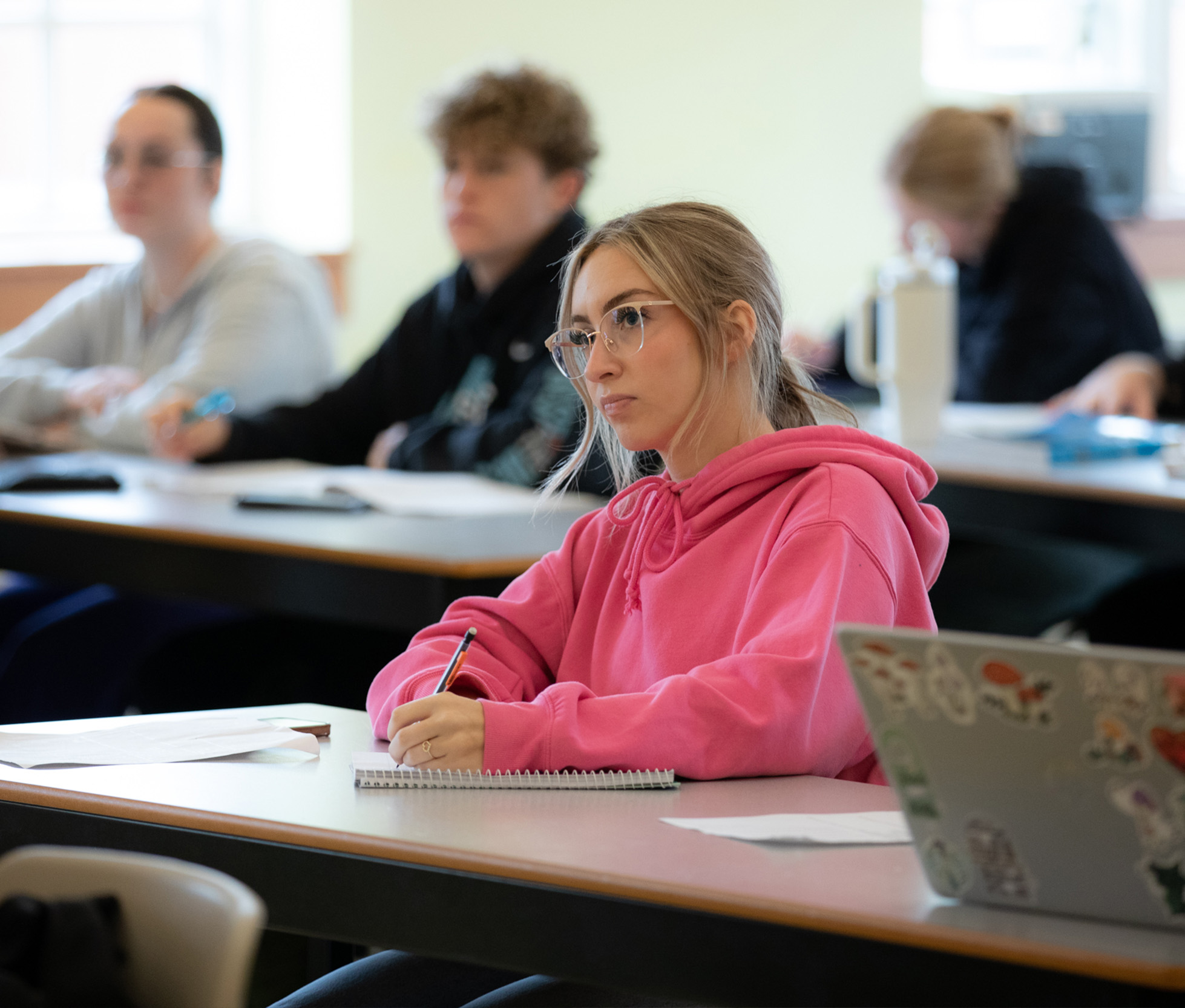 A female student wearing a pink sweater takes notes in class