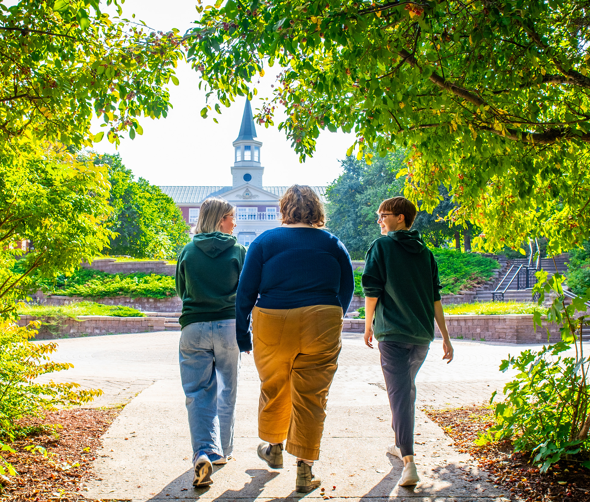 Three students walk through the trees toward STU's lower courtyard