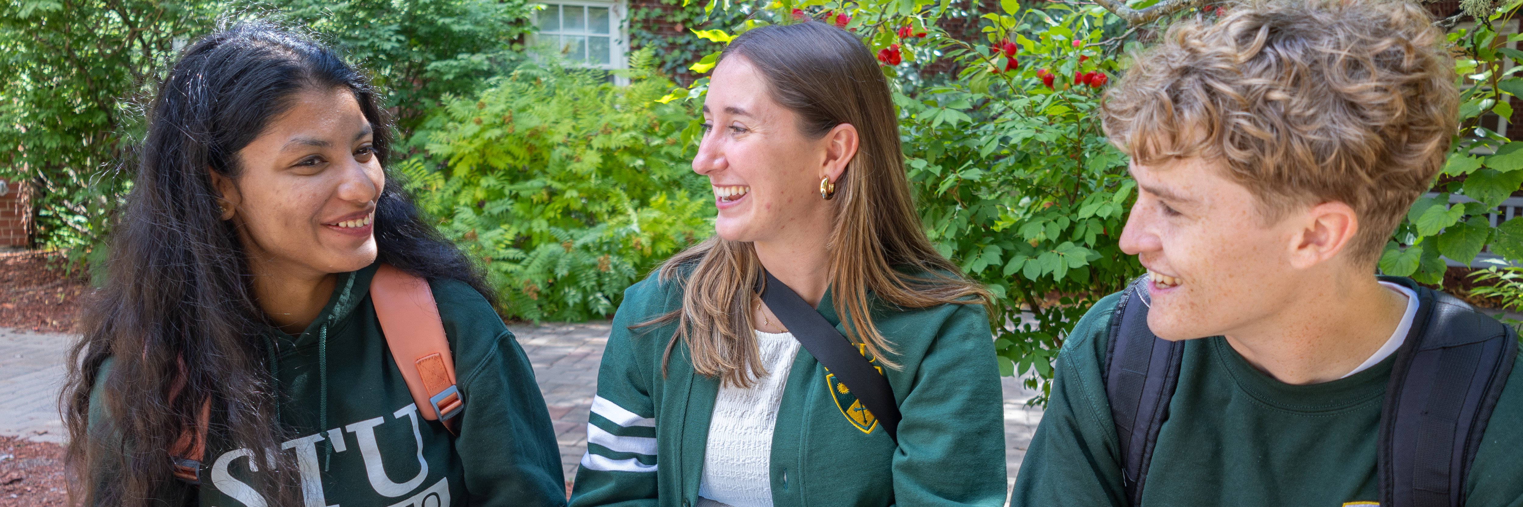 Two female students and a male student sit together chatting on campus in the fall