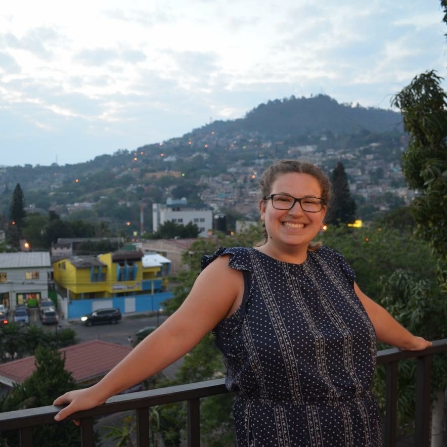 A woman with glasses and braided hair wearing a navy patterned dress leans against a railing in front of a city