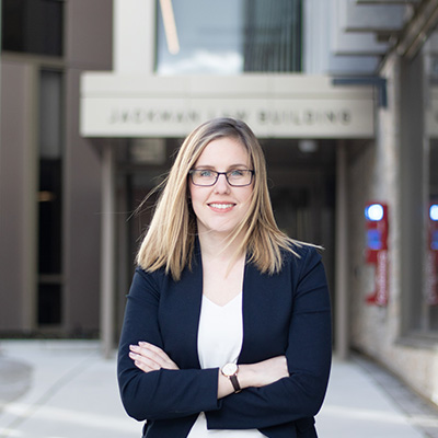 A woman wearing a black blazer and white shirt stands with her arms crossed in front of the entrance to an office building