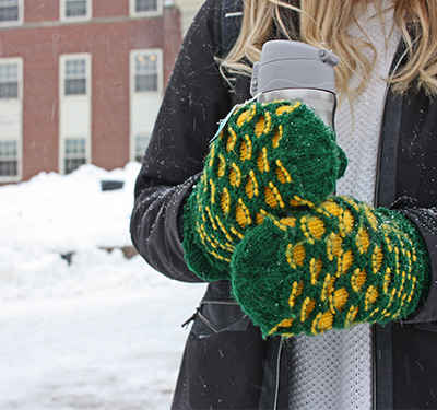 Student holding flask with green and yellow mittens outside in snowy weather