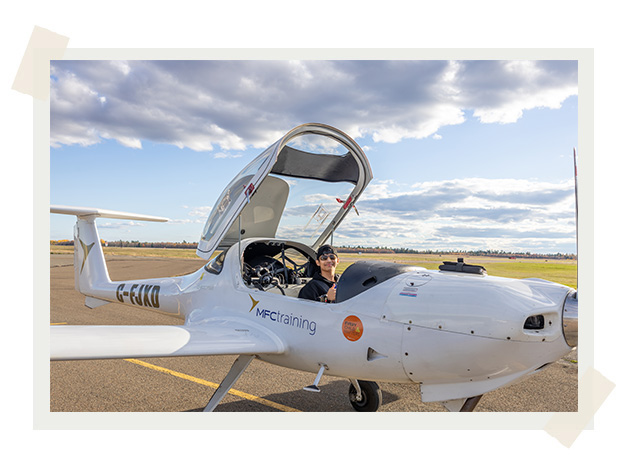 A male student sits in a small plane with the top open, giving a thumbs up.