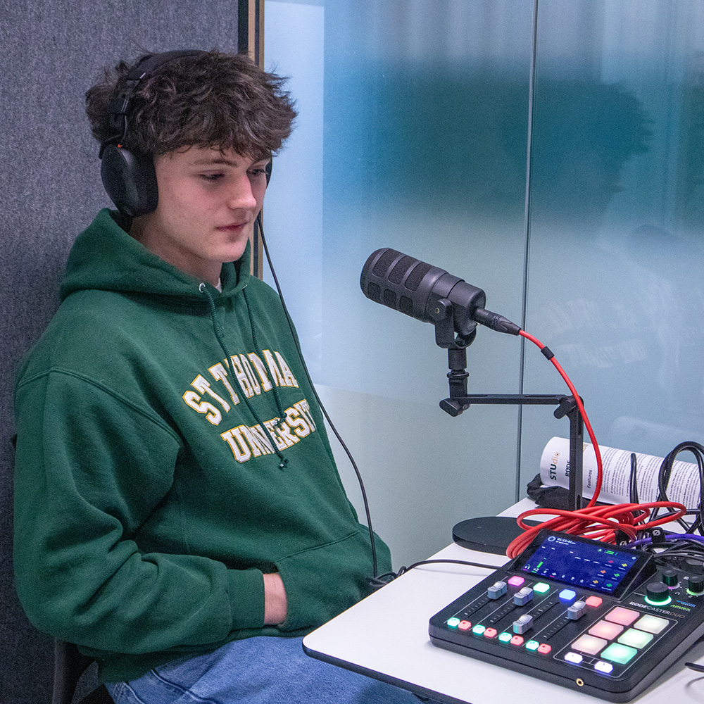 A male student wearing a St. Thomas sweater sits in the podcast booth wearing headphones