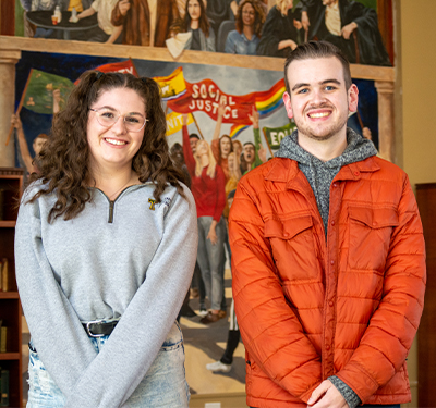 Two students standing in the Great Hall with social justice poster painting in background