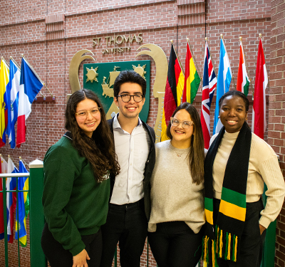 Four international students smiling next to wall of flags in James Dunn Hall at STU