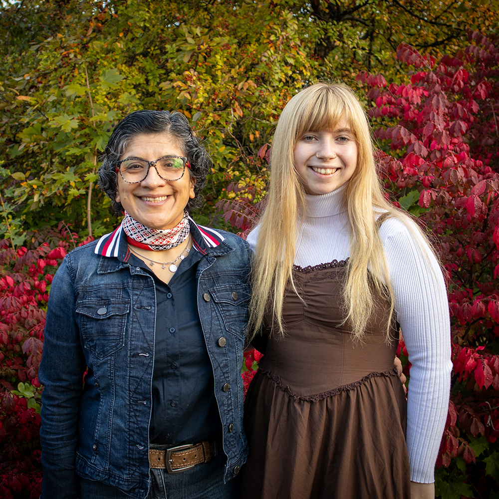 A female professor and female student stand side by side in front of fall leaves on campus