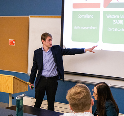 Male student presenter standing in classroom sharing findings to peers and professor