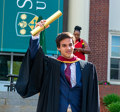 Graduating student in gown holding diploma in front of George Martin Hall