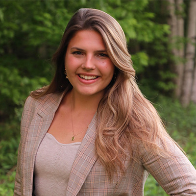 A woman wearing a tan blazer and top standing in front of greenery