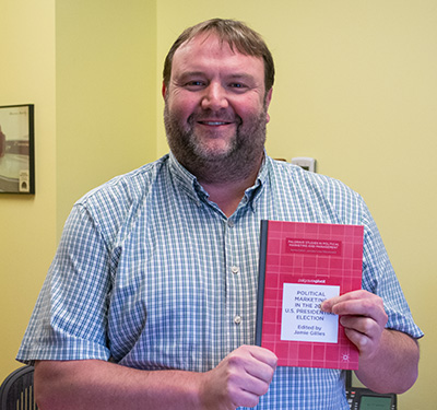 Dr. Jamie Gillies stands in his office holding his book.