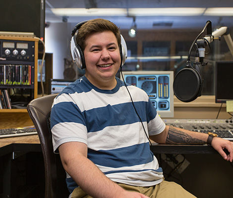 A male student wearing headphones sits in front of a microphone in a recording studio.