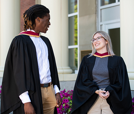 Two students in graduation gowns having a conversation in front of GMH doorway