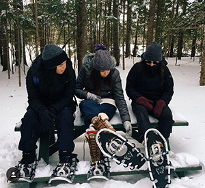 Three students wearing snowshoes sitting in the forest. 